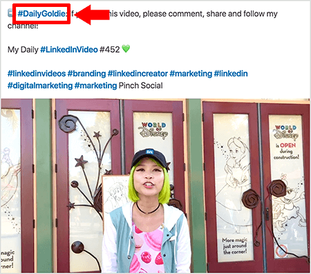 זהו צילום מסך שממחיש כיצד גולדי צ'אן משתמשת בהאשטגים בטקסט של הודעות הווידיאו שלה בלינקדאין. הסברים אדומים מצביעים על ההאשטג #DailyGoldie בטקסט, שהוא ייחודי לפוסטי הווידיאו שלה ועוזר לה לעקוב אחר שיתופים. הפוסט כולל גם hashtags רלוונטיים אחרים המסייעים לאנשים למצוא את הסרטון שלה, כולל #LinkedInVideo. בתמונת הווידיאו גולדי עומדת מול כמה דלתות בתצוגה של עולם דיסני. היא אישה אסייתית עם שיער ירוק. היא חובשת כיפה שחורה של לינקדאין, שרשרת קולר שחורה, חולצת הדפסת מקרון ורודה וז'קט כחול לבן.